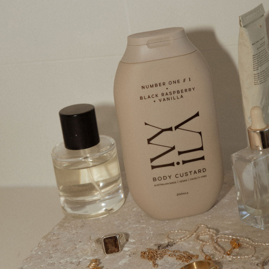 Body custard bottle with other skincare products and jewelry on a textured surface.