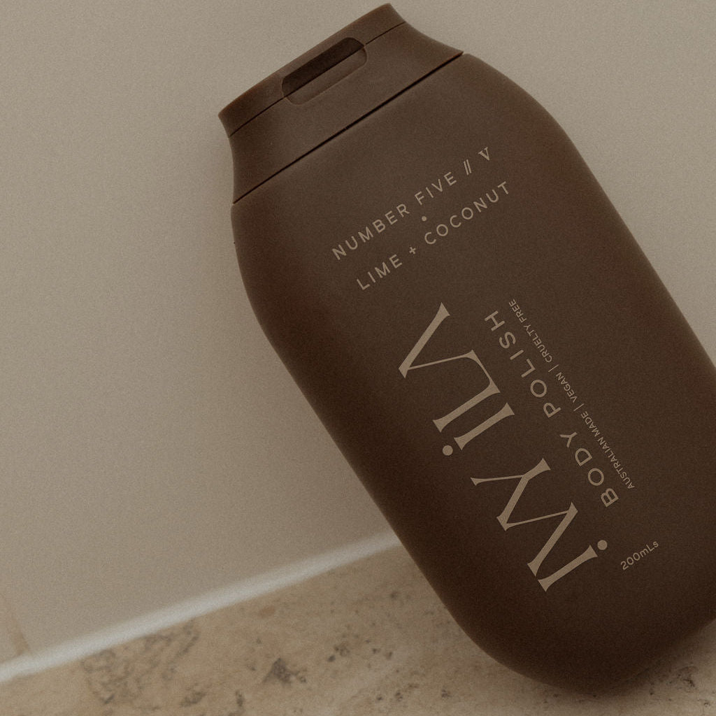 Brown bottle of IMWIL body polish on a beige floor against a beige wall