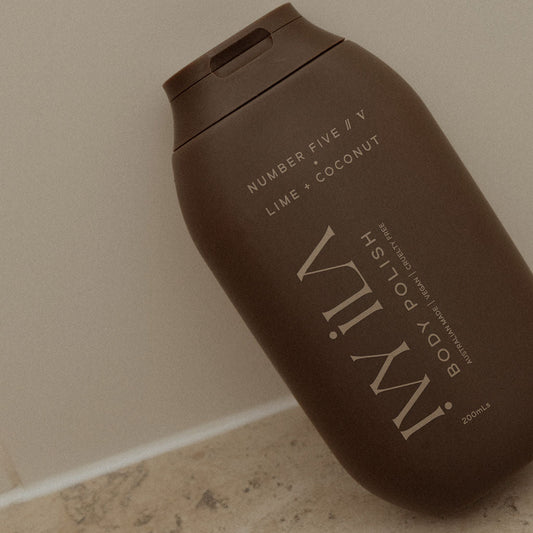 Brown bottle of IMWIL body polish on a beige floor against a beige wall