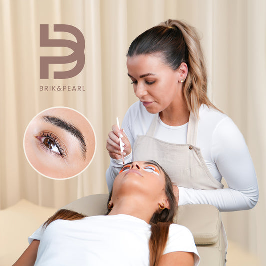 A brow specialist applies makeup to a client's eyebrow while the client lies down, with a circular inset showing a close-up of the eyebrow. The BriK&Pearl logo is visible in the top left.