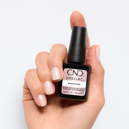 A hand holding a bottle of CND Shellac nail dip powder in the shade Bompayante. The nails are painted with a glossy, pale pink nail polish, and the hand is set against a clean, light gray background.