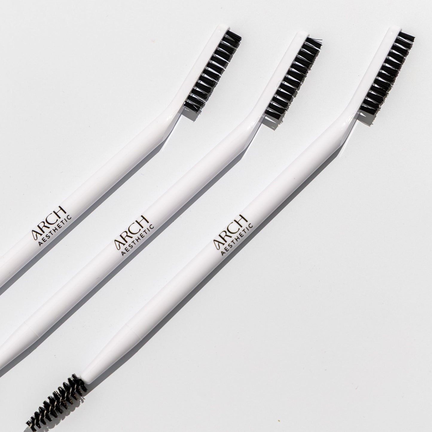 Three white eyebrow brushes with black bristles on a light gray background, branded 'ARCH'.