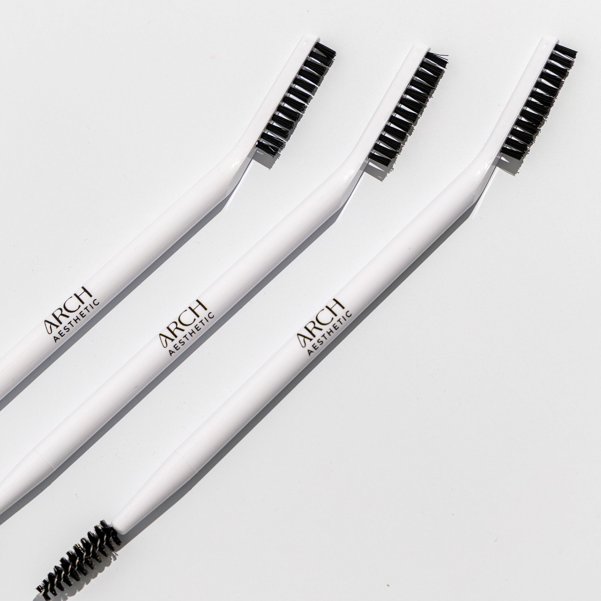 Three white eyebrow brushes with black bristles on a light gray background, branded 'ARCH'.