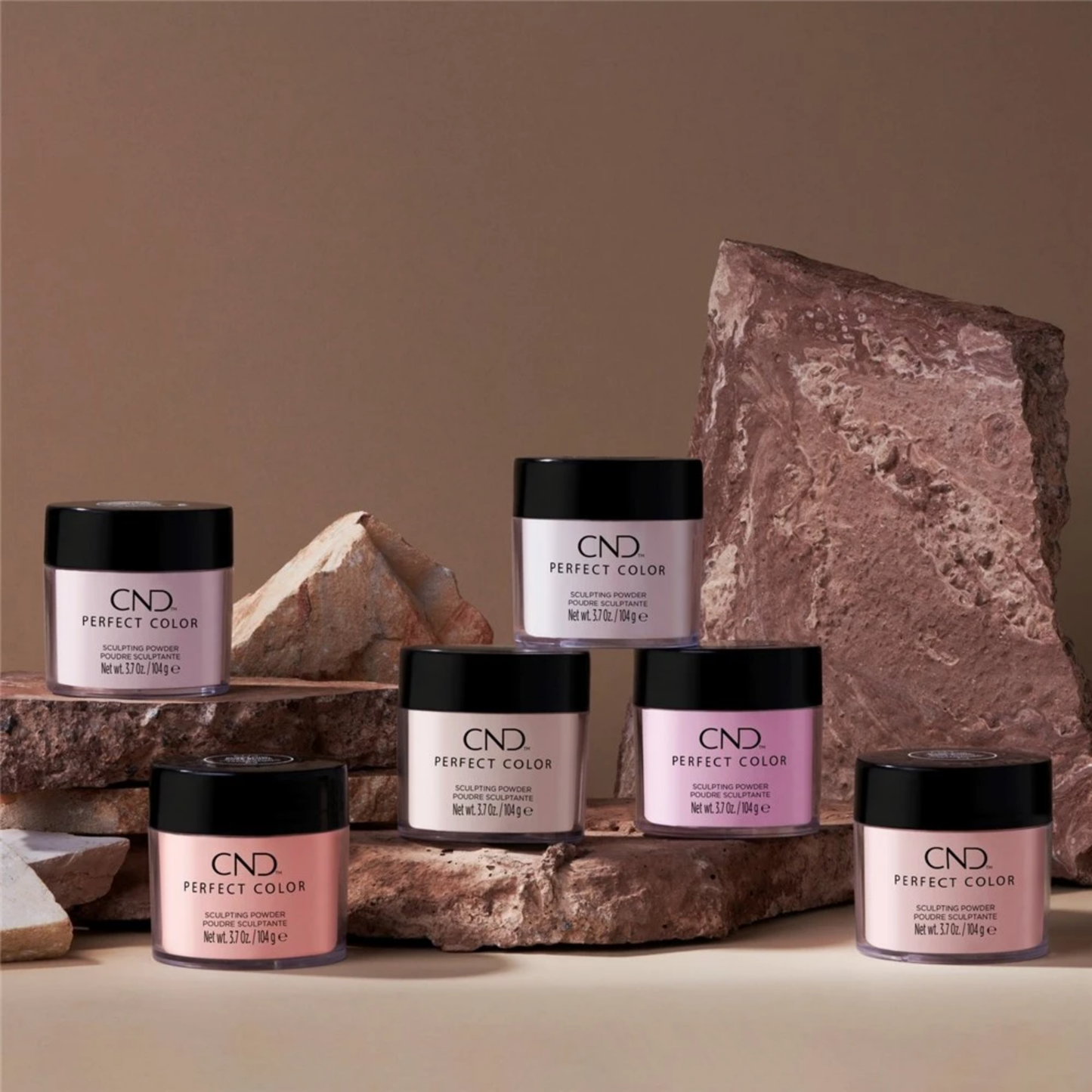 Six jars of CND Perfect Color Pressed Powder arranged on and around rough stone blocks against a warm beige background. The jars contain light pink, lavender, peach, and off-white powders, with black lids and white labels.