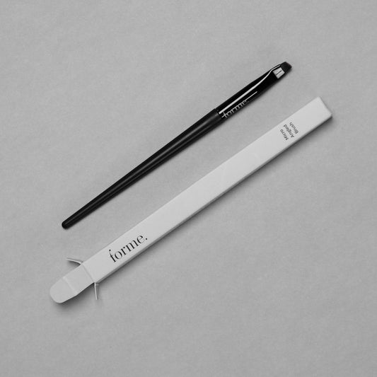 A micro angled brush with a black handle and silver bristles, placed next to its white packaging with the brand 'forme' printed on it.