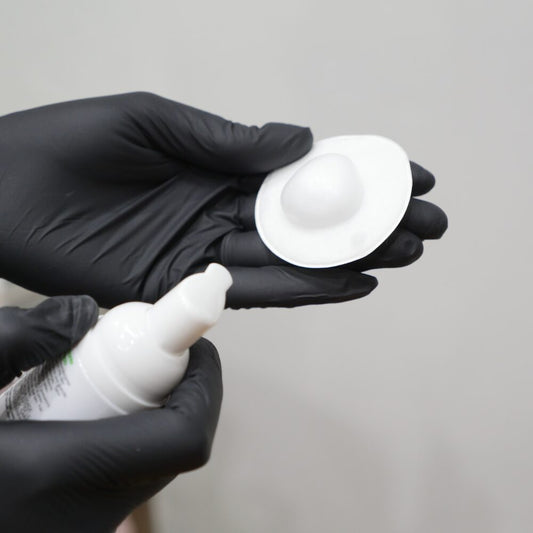 Person wearing black gloves holding a white container and applicator against a gray background