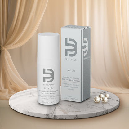 A white cylindrical bottle and matching gray-and-white product box for BrikPearl Lash Life intensive conditioning hair for lashes and brows, displayed on a round marble pedestal with draped beige fabric and pearl-like beads in the background.