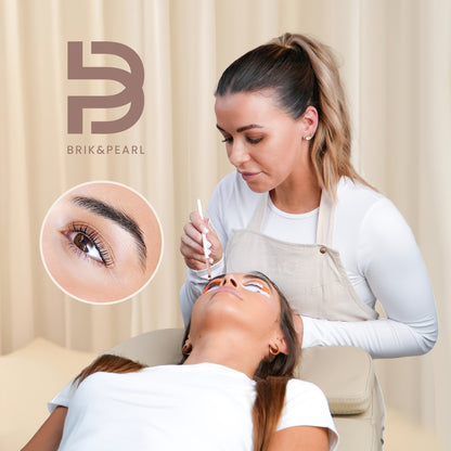 A brow specialist applies makeup to a client's eyebrow while the client lies down, with a circular inset showing a close-up of the eyebrow. The BriK&Pearl logo is visible in the top left.