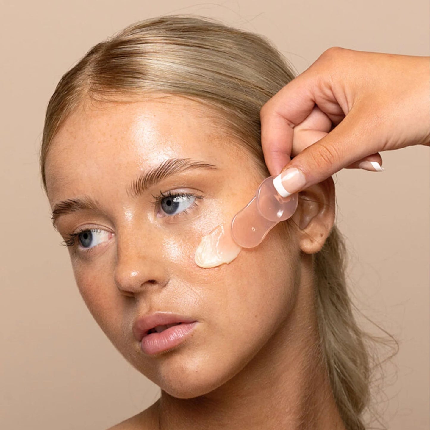 Woman having cream applied to her face by a hand against a beige background