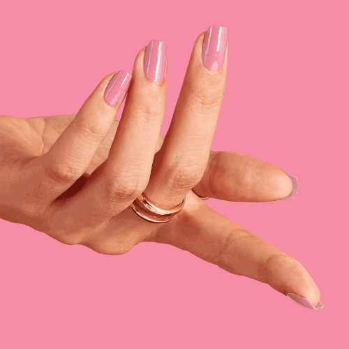 A close-up of a hand with long, glossy pink manicured nails wearing two slim rose gold rings, set against a solid pink background.