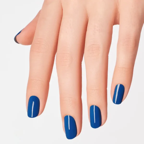 A close-up of a hand with neatly manicured nails painted in a solid medium blue polish, set against a clean, light gray background.